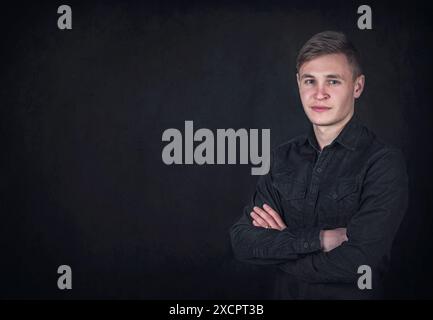 Porträt eines selbstbewussten jungen Mannes, hält die Arme gefaltet vor einem schwarzen Wandhintergrund mit Kopierraum. Erfolgreicher männlicher Student mit entschlossenem Gesichtsausdruck Stockfoto