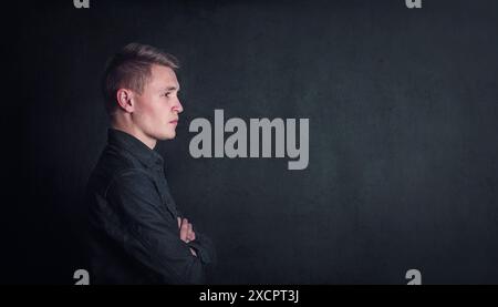 Nachdenkliches Porträt junger Mann mit Seitenansicht, hält die Arme gegen einen dunklen Wandhintergrund gefaltet und mit Kopierraum. Nachdenklicher männlicher Student mit entschlossenem Gesicht e Stockfoto