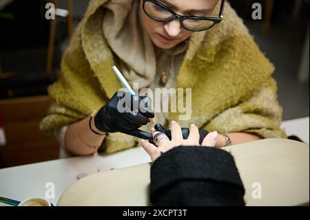 Nahaufnahme eines erfahrenen Nagelkünstlers, der in einem Schönheitssalon komplizierte Designs auf die Nägel eines Kunden malt und Präzision und Kunstfertigkeit unterstreicht. Stockfoto