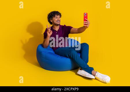 Foto in voller Größe des netten jungen Mannes Selfie Foto V-Zeichen Sit Sitzsack Gerät tragen trendiges violettes Outfit isoliert auf gelbem Hintergrund Stockfoto