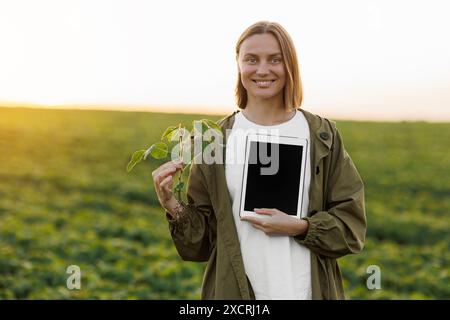 Die lächelnde Agronomin hält ein digitales Tablet mit Mockup-Bildschirm und Sojapflanze im Feld, untersucht, überprüft, schaut in die Kamera. Smart Farming so Stockfoto