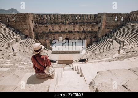 Eine Frau mit Hut sitzt auf den steilen Steinen im berühmten antiken Theater von Aspendos in der Nähe von Antalya in der Türkei an einem sonnigen Tag mit blauem Himmel. Stockfoto