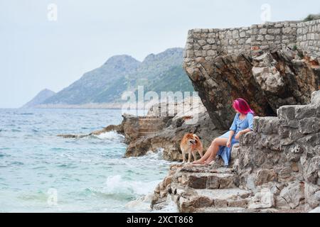 Eine rosafarbene Frau und ein Labrador Retriever teilen sich einen ruhigen Moment auf alten Stufen am Meer, während das Meer neben ihnen flüstert Stockfoto