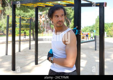 Ein Mann mit einem blauen Armband steht vor einer Metallkonstruktion. Er trägt ein weißes Tanktop und schwarze Shorts Stockfoto