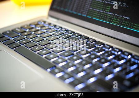 Aufnahme der beleuchteten Tastatur eines modernen Laptops mit sichtbarem Programmiercode auf dem Bildschirm, der fortschrittliche Technologie, Hacking, Data Brach und ddos betont Stockfoto