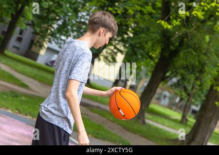 Ein junger Teenager in einem grauen Hemd und schwarzen Shorts hält einen Basketball in der rechten Hand. Er steht auf einem gepflasterten Außenplatz in einem Wohngebiet. T Stockfoto