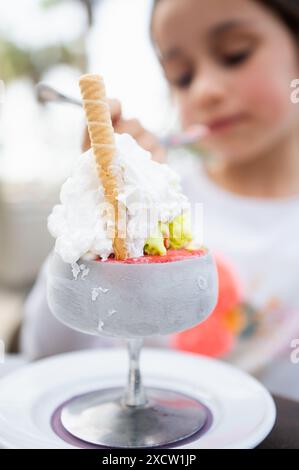 Nahaufnahme eines Kindes, das einen farbenfrohen Eisbecher mit Schlagsahne und einem Waffelstab genießt. Ein wunderbarer Genuss an einem sonnigen Tag. Stockfoto