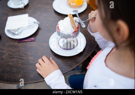 Ein Kind genießt in einem Café im Freien einen köstlichen Eisbecher mit Schlagsahne und einer Waffel. Die Szene fängt einen Moment des Glücks und der Liebe ein Stockfoto