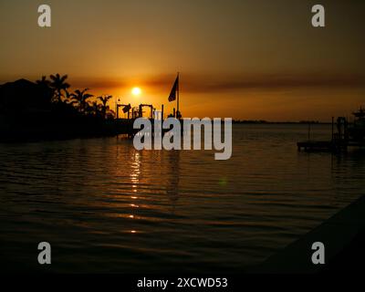 Sonnenuntergang Silhouette der US-Flagge mit Blick auf NW Boca Ciega Bay in der Nähe der Brücke am Treasure Island Causeway. Palmen und Bootssteg rechts. Sonne nahe dem Horizont Stockfoto