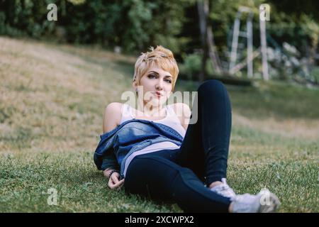 Junge Frau mit kurzen blonden Haaren liegt auf dem Gras und genießt einen sonnigen Tag im Park. Sie trägt legere Kleidung und hat eine entspannte und ca. Stockfoto