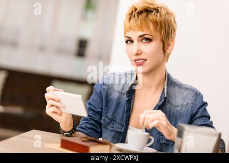 Eine junge Frau mit kurzen blonden Haaren genießt ihren Kaffee in einem gemütlichen Café und macht ein Foto davon auf ihrem Handy. Sie sieht entspannt und zufrieden aus und strahlt einen Beifall aus Stockfoto