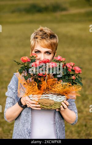 Die junge Frau hält einen Korb mit orangefarbenen Rosen vor ihrem Gesicht, umgeben von einem üppigen grünen Feld. Sie genießt die Schönheit der Natur und die s Stockfoto