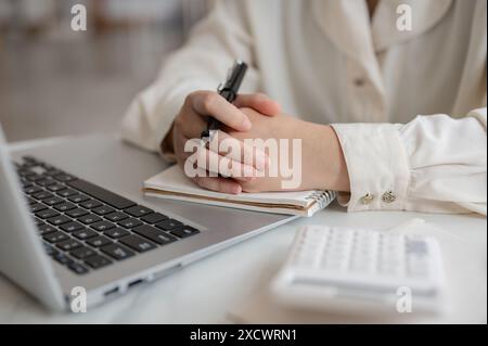 Ein Nahbild einer seriösen Geschäftsfrau, die mit aneinander gefalteten Händen an ihrem Schreibtisch sitzt, ein Laptop auf dem Schreibtisch. Stockfoto
