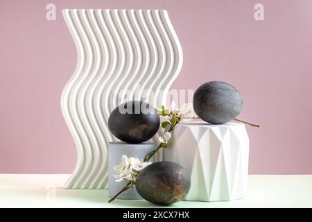 Ostereier in schwarzer Farbe und Kirschblütenzweig auf dekorativen weißen Podesten auf hellem Fliederhintergrund. Stillleben, Kopierraum. Stockfoto