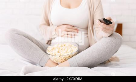 Schwangere Frau, die mit Popcorn und Fernbedienung auf dem Bett sitzt Stockfoto