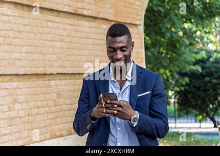 Afroamerikanischer Geschäftsmann in elegantem Anzug, der mit seinem Smartphone läuft und eine Nachricht sendet. Junger professioneller Manager lächelt, während er seine benutzt Stockfoto