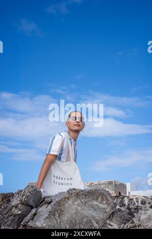 Ein Mann mit Sonnenbrille und gestreiftem Hemd steht auf einem Felsvorsprung mit blauem Himmel und weißen Wolken im Hintergrund. Stockfoto