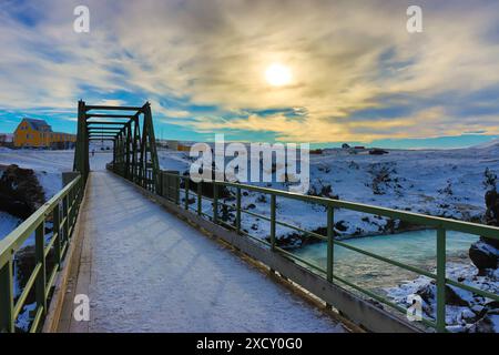 Winterlandschaft einer schneebedeckten Brücke, die über einen Wasserfallbach in Island führt. Ein Sonnenuntergangshimmel mit Wolken. Stockfoto