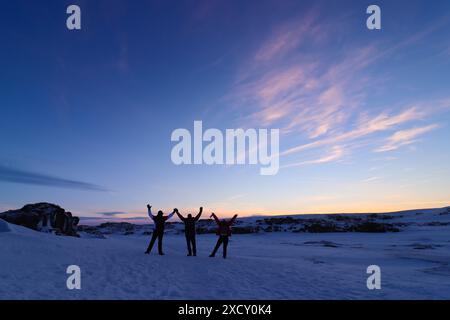 Sonnenuntergang in Island mit drei Freunden, die Spaß im Schnee haben. Drei Silhouetten von Menschen mit erhobenen Armen in einer Winterlandschaft. Stockfoto
