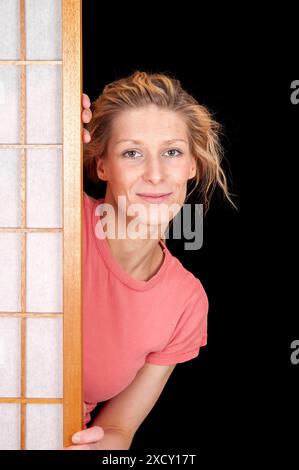Oberkörperporträt einer jungen blonden Frau, die hinter einem Bildschirm steht und seitlich vor schwarzem Hintergrund in die Kamera blickt. Stockfoto