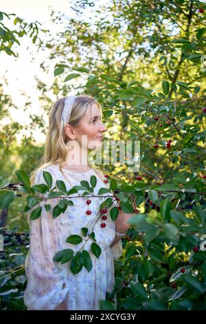 Ein Porträt einer jungen blonden Frau in einem pastellfarbenen Chiffonkleid zwischen den Zweigen einer Reifen Kirsche Stockfoto