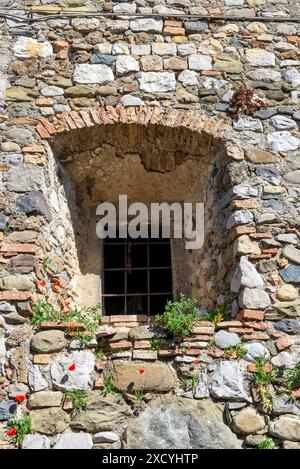 Italienisches Fenster in einem alten Gebäude mit dicken Wänden und umgeben von wilden Blumen, die aus den Rissen im Stein wachsen Stockfoto