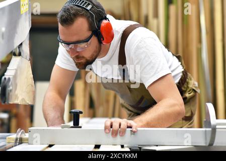 Junger Zimmermann in Arbeitskleidung arbeitet in der Schreinerei an einer Schleifmaschine - Arbeitskleidung mit Gehörschutz Stockfoto