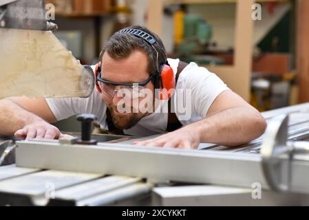 Junger Zimmermann in Arbeitskleidung arbeitet in der Schreinerei an einer Schleifmaschine - Arbeitskleidung mit Gehörschutz Stockfoto