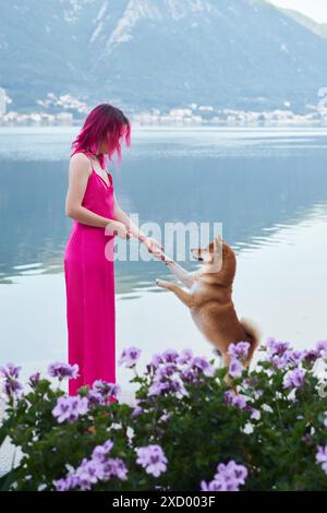 Eine spielerische Interaktion entfaltet sich, wenn eine Frau in einem rosa Kleid ihrem Shiba Inu Hund beibringt, am See zu stehen, umgeben von violetten Blumen Stockfoto