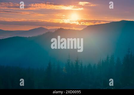 Ein malerischer Blick auf eine Bergkette bei Sonnenaufgang, mit einer trüben Nebelschicht, die die bewaldete Landschaft bedeckt. Die Sonne blickt über den Horizont und strahlt ein warmes Licht auf die Wolken. Stockfoto