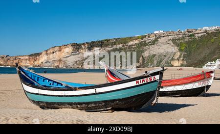 Farbenfrohe traditionelle Fischerboote am Sandstrand in Nazare, Portugal. Farbenfrohe Fischerboote ruhen am Sandstrand mit Klippen und klarem blauem Himmel. Stockfoto