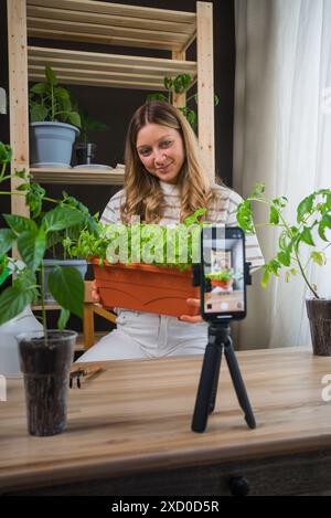 Frau, die Pflanzgefäße mit grünen Kräutern hält, während sie mit dem Smartphone gefilmt wird. Sie sitzt in gemütlicher Gartenanlage, umgeben von verschiedenen Topfpflanzen, die Freude an der Gartenarbeit in Innenräumen zum Ausdruck bringen und sie über digitale Medien teilen. Stockfoto