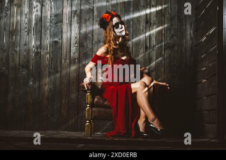 Frau mit Calavera-Make-up, die den Tag der Toten symbolisiert, sitzt elegant auf einem Vintage-Stuhl. Sie trägt ein markantes rotes Kleid und einen floralen Kopfschmuck mit roten und schwarzen Rosen vor dunklem Holzhintergrund. Stockfoto