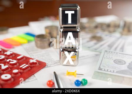 Ein Stapel Würfel mit dem Wort "Tax" darauf. Die Würfel stehen auf einem Stapel Münzen und einem Stapel Dollarscheine Stockfoto