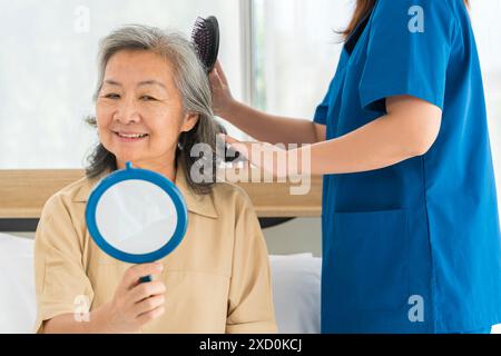 Eine Frau sitzt in einem Bett mit einem Spiegel vor sich. Eine Frau in blauer Uniform steht hinter ihr und bürstet sich die Haare. Stockfoto