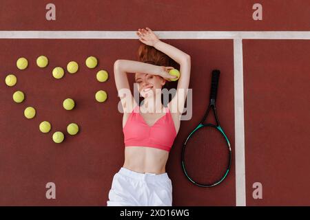 Junge Tennisspielerin liegt auf dem Tennisplatz, Tennisbälle in Form eines Herzens, Blick von oben, Tennismeisterschaft Stockfoto