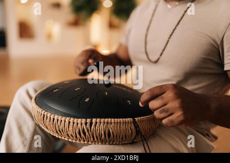 Nahaufnahme eines nicht erkennbaren schwarzen Mannes in Trance, der es genießt, mit Sticks auf Glucophon zu spielen, sitzend im Kreuz auf dem Boden zu Hause mit gemütlichem Interieur. Stockfoto