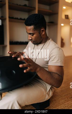 Nahaufnahme eines fokussierten afrikanischen jungen Mannes in Trance, der mit Händen auf einer Stahlzungentrommel auf dem Boden zu Hause mit gemütlicher Innenausstattung sitzt. Stockfoto