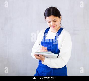 Interessierte Mechanikerin in blauen Overalls, die die Reihenfolge aufschreibt Stockfoto