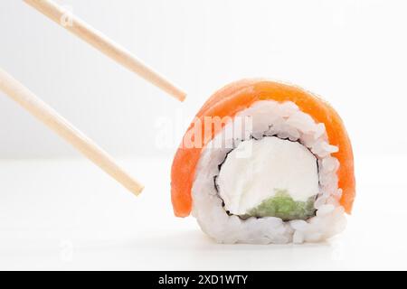 Nahaufnahme eines runden Sushi-Brötchens mit frischem Lachs, der von Stäbchen gehalten wird. Das Sushi-Brötchen enthält Lachs, Reis, Frischkäse und Avocado. Japanische Küche Stockfoto