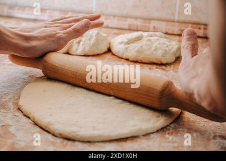 Hände, die Teig mit einem Holzstift ausrollen. Zwei Teigkugeln befinden sich im Hintergrund auf einer bemehlten Oberfläche. Stockfoto