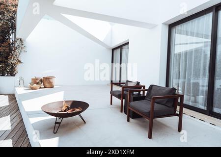 Eine moderne Außenterrasse mit minimalistischem Design, zwei Holzstühlen mit dunklen Kissen, eine Feuerstelle aus Metall und Topfpflanzen vor weißen Wänden. Stockfoto