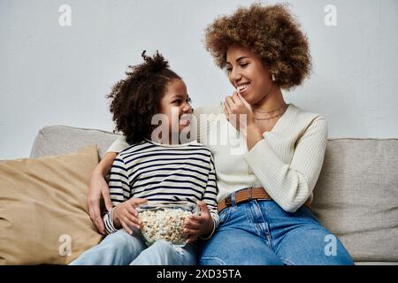 Glückliche afroamerikanische Mutter und Tochter sitzen auf einer Couch und genießen gemeinsam einen Snack Popcorn. Stockfoto