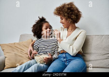 Eine afroamerikanische Mutter und Tochter sitzen glücklich auf einer Couch und genießen Popcorn zusammen. Stockfoto