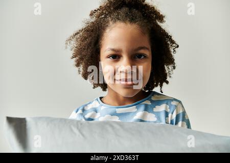 Ein junges Mädchen, in Pyjamas gekleidet, versteckt ihr Gesicht verspielt hinter einem Kissen und bringt Freude und Lachen in die Szene. Stockfoto