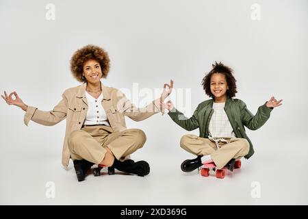 Lockige afroamerikanische Mutter und Tochter in stilvollen Outfits, sitzen auf Skateboards und üben gemeinsam Yoga auf grauem Hintergrund. Stockfoto