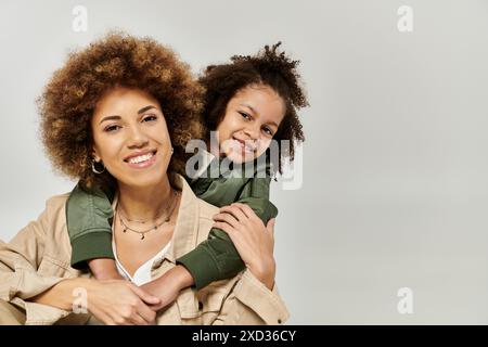 Stilvolle afroamerikanische Mutter mit lockigen Afro-Haaren trägt ihre Tochter auf ihren Schultern vor grauem Hintergrund. Stockfoto