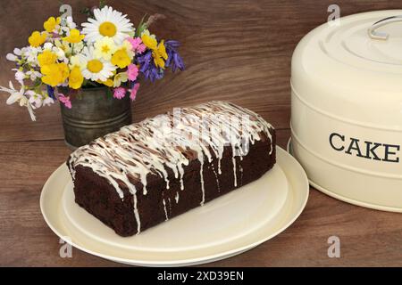 Schokoladensplitterkuchen mit Zuckerguss auf Metallschale mit Deckel auf rustikalem Holzhintergrund mit Frühlingsblumen und Wildblumen. Hausgemachte köstliche Leckerbissen. Stockfoto