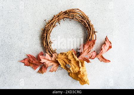 Ein rustikaler Herbstkranz aus miteinander verflochtenen Zweigen, verziert mit verstreuten getrockneten Blättern in Rot-, Gelb- und Brauntönen, präsentiert auf einem grauen Text Stockfoto