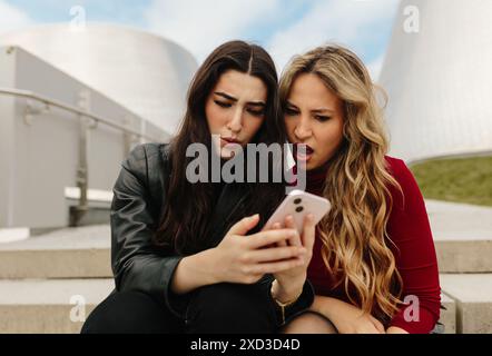Zwei junge Frauen sitzen auf Stufen vor der Tür und teilen einen schockierenden oder überraschenden Moment, während sie auf ein Smartphone schauen Stockfoto